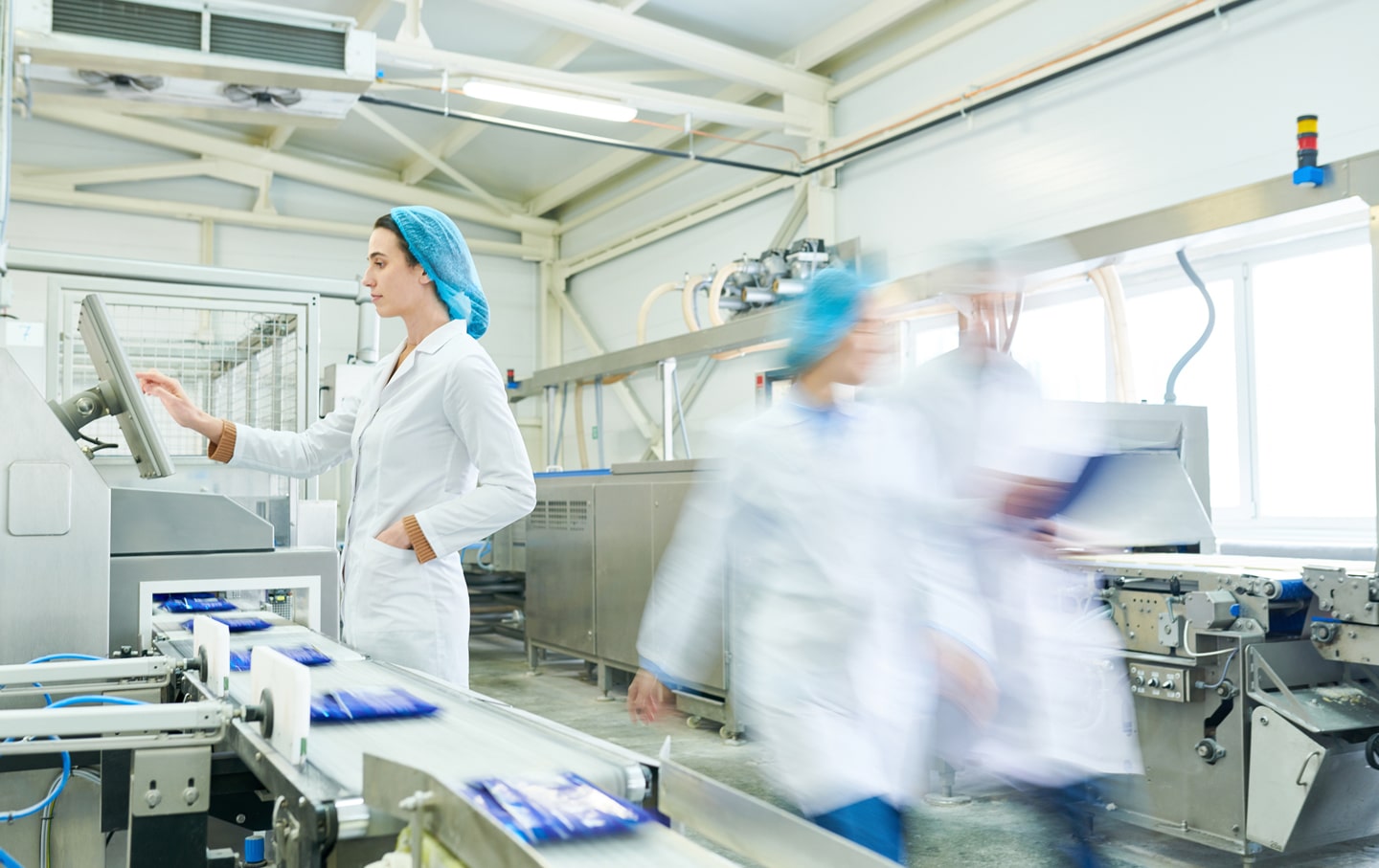 Female worker operating touchscreen controls on a food production line, with packaged products on a conveyor and other staff in motion in a clean, modern processing facility.