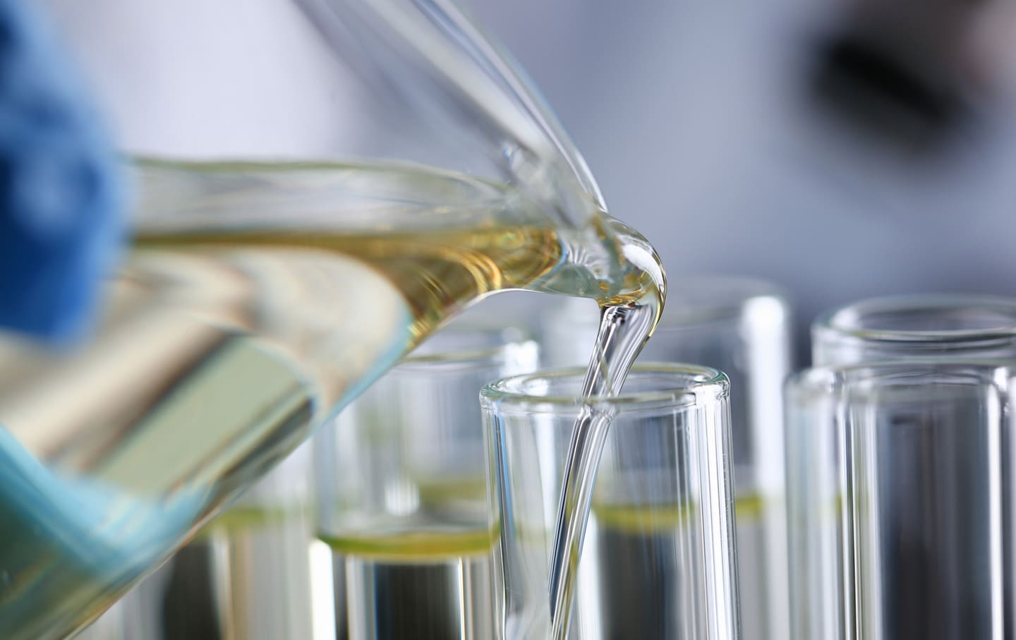 Close-up of a clear liquid being poured from a laboratory beaker into a test tube, representing chemical formulation and quality control in a hygiene product lab.