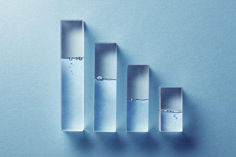 Conceptual bar graph made of transparent glass columns filled with water at varying levels, symbolising reduction or efficiency metrics on a blue background.