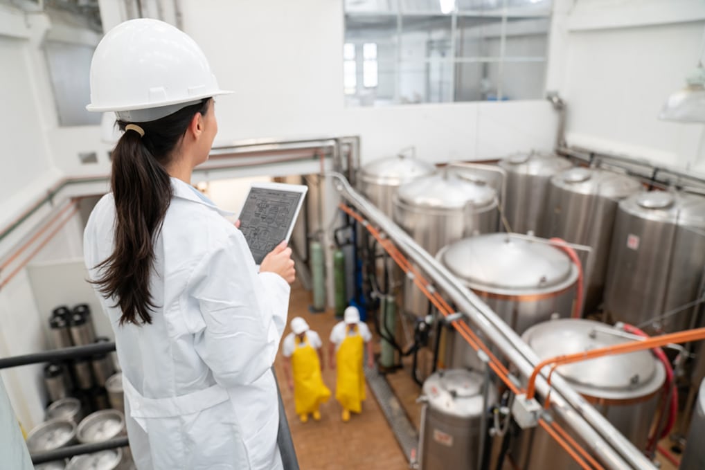 Female supervisor in a hard hat and lab coat holding a tablet, overseeing beverage processing operations with large stainless steel tanks and workers in yellow aprons.