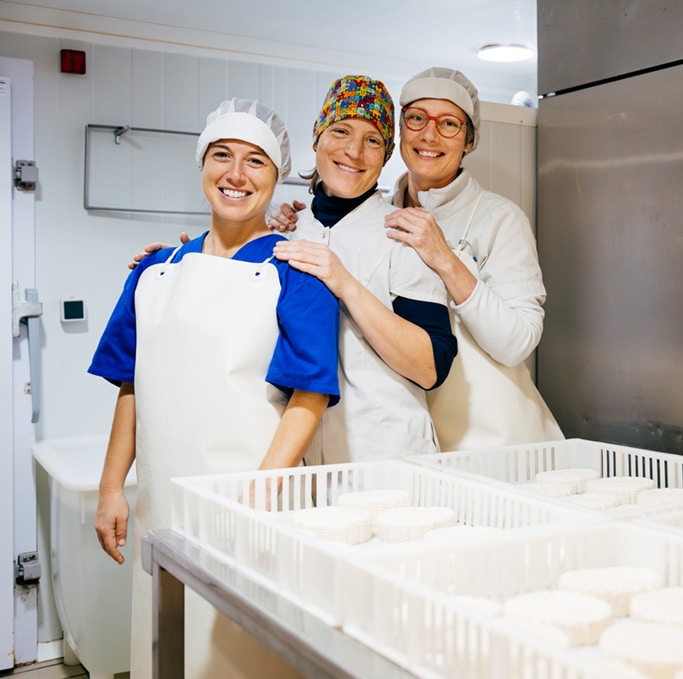 Three smiling workers in protective clothing standing together in a cheesemaking facility, with trays of artisanal cheese rounds in the foreground.