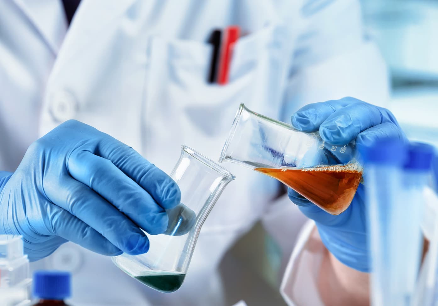 Scientist wearing blue gloves pouring coloured liquids between laboratory glassware during chemical formulation product testing.