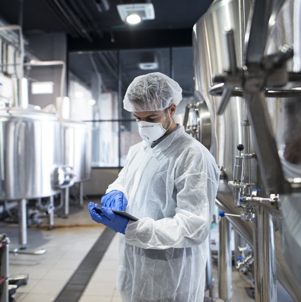 Food industry worker in full hygiene PPE inspecting equipment in a stainless steel processing facility.