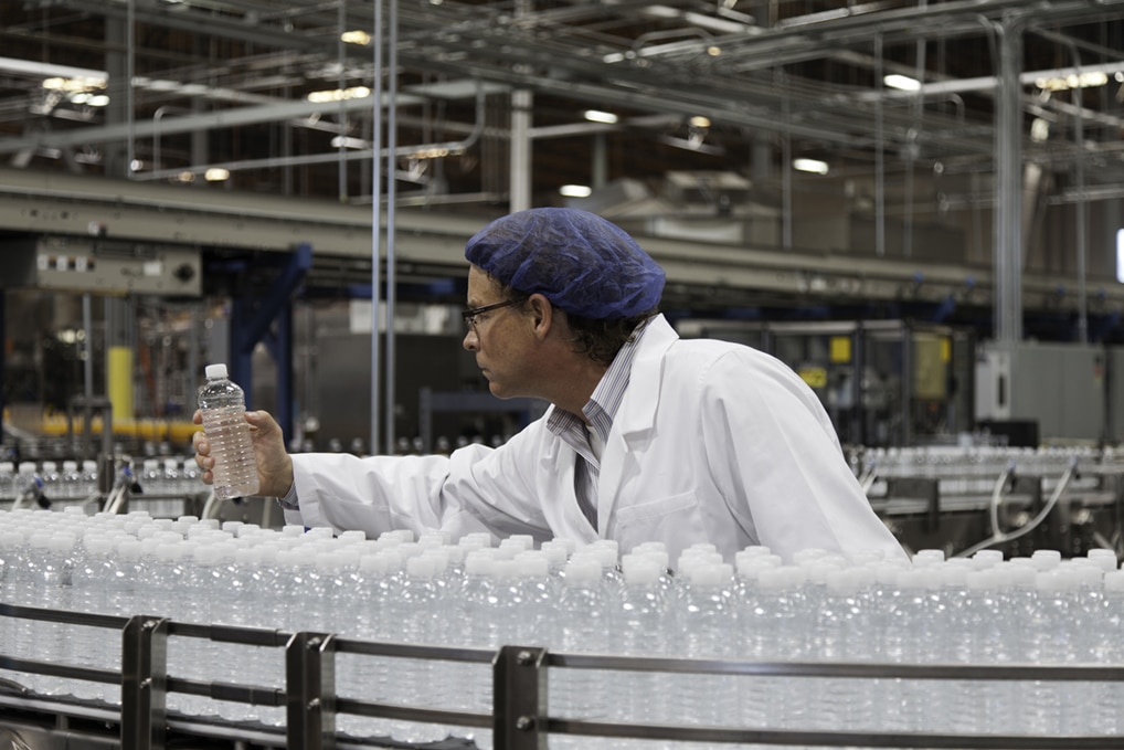 Quality control inspector examining a bottle of water on a bottling line in a beverage production facility, surrounded by rows of identical sealed bottles.