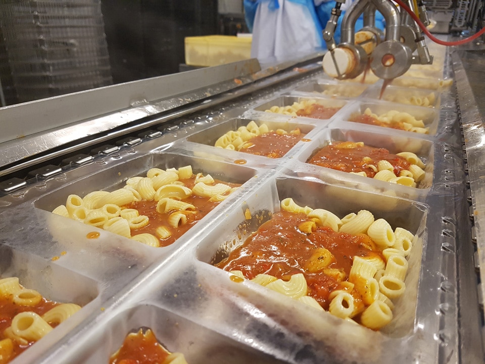 Automated filling system dispensing tomato sauce over cooked pasta in plastic trays on a ready meal production line.