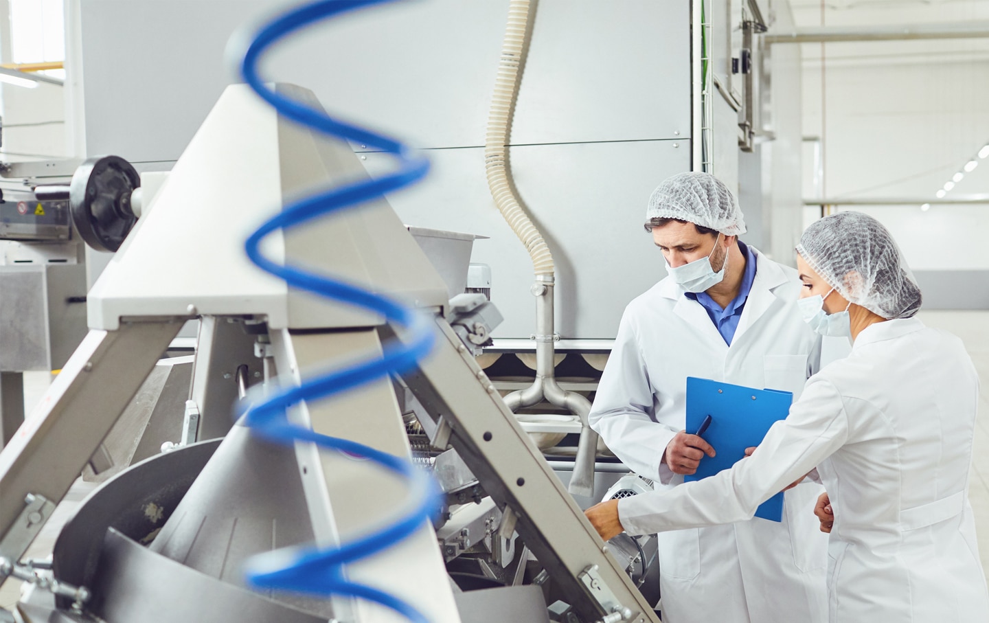http://Two%20food%20production%20workers%20in%20lab%20coats,%20hairnets%20and%20face%20masks%20inspecting%20industrial%20processing%20equipment%20with%20a%20clipboard%20in%20a%20clean,%20modern%20facility.