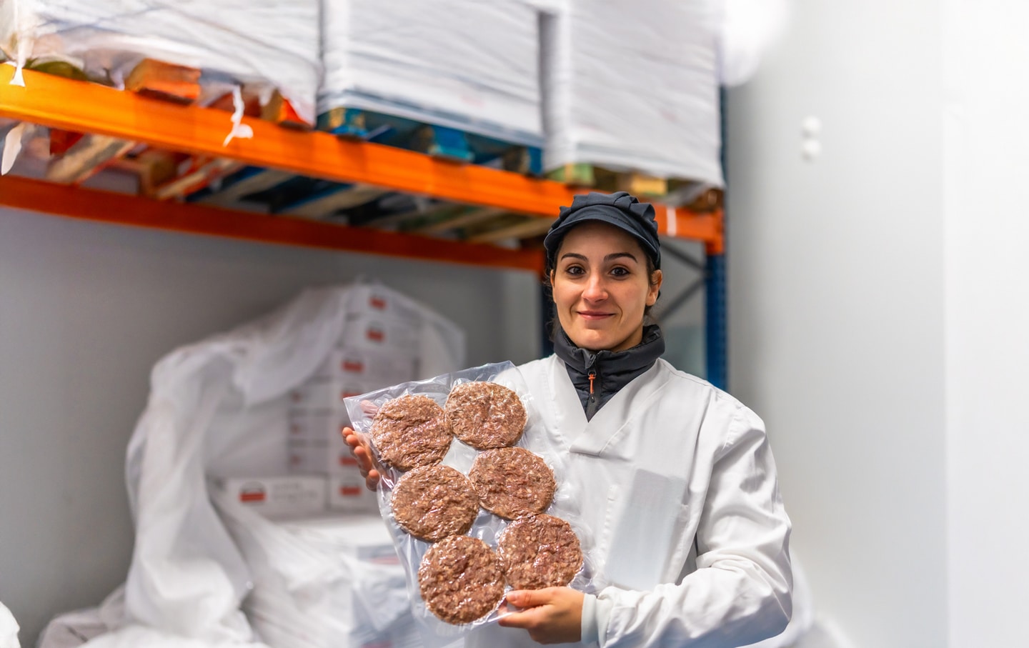 http://Food%20factory%20worker%20in%20protective%20clothing%20holding%20a%20vacuum-sealed%20pack%20of%20frozen%20beef%20burgers%20inside%20a%20cold%20storage%20facility.